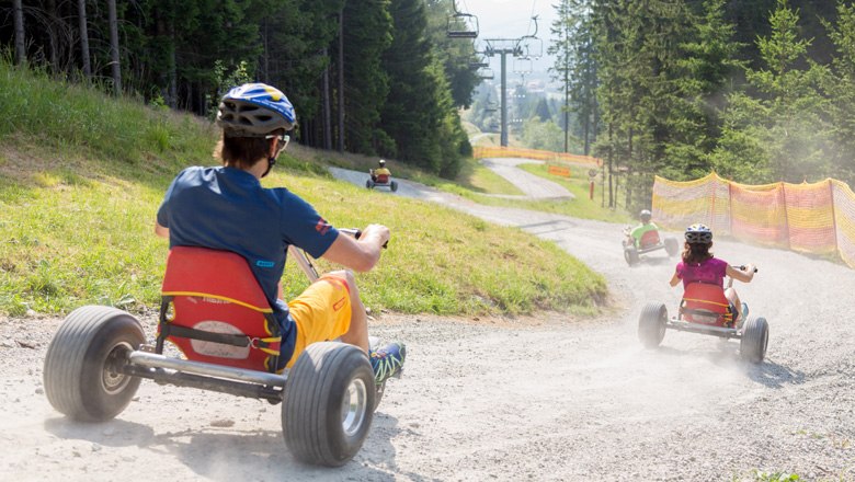 People ride down a gravel path on mountain carts, surrounded by trees and a ski lift.