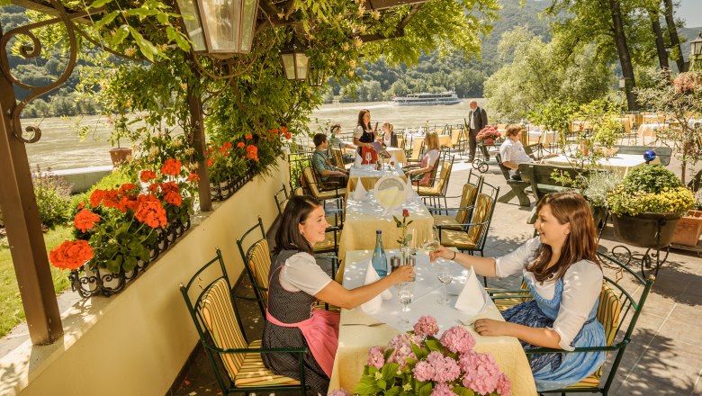 People sitting in a garden restaurant with a view of a river, surrounded by flowers and trees.