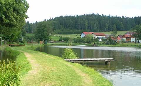 A quiet lake with a jetty, surrounded by green countryside and houses in the background.