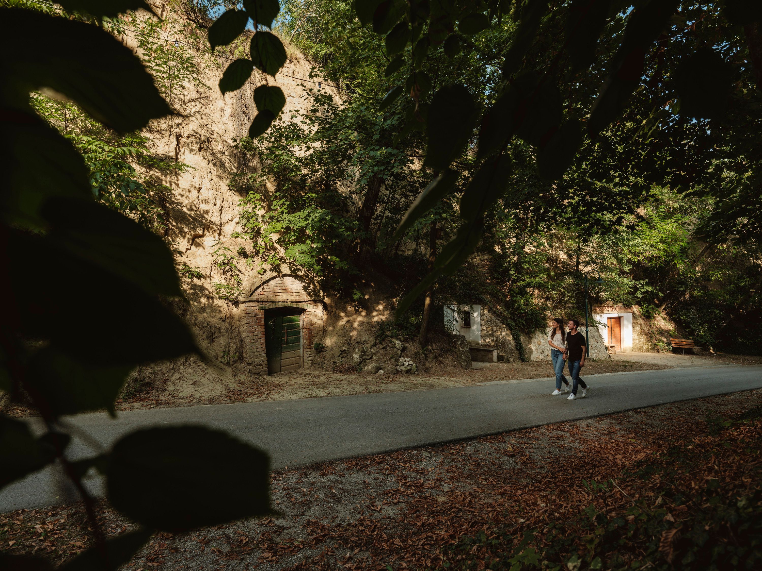 Two people are walking along a road in a green, wooded area with wine cellars in a rock face.