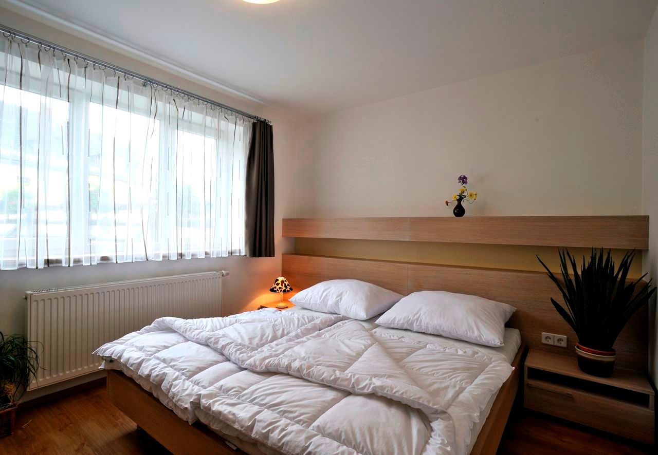 Modern bedroom with double bed, light wooden furniture and large windows with white curtains.