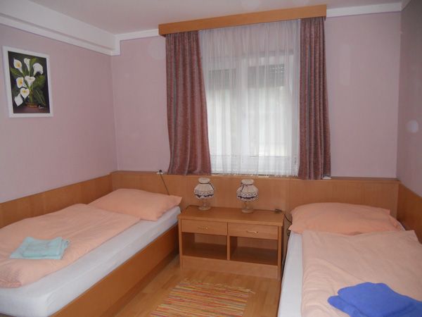 A simply furnished room with two single beds, a window with curtains and a small table with two lamps.