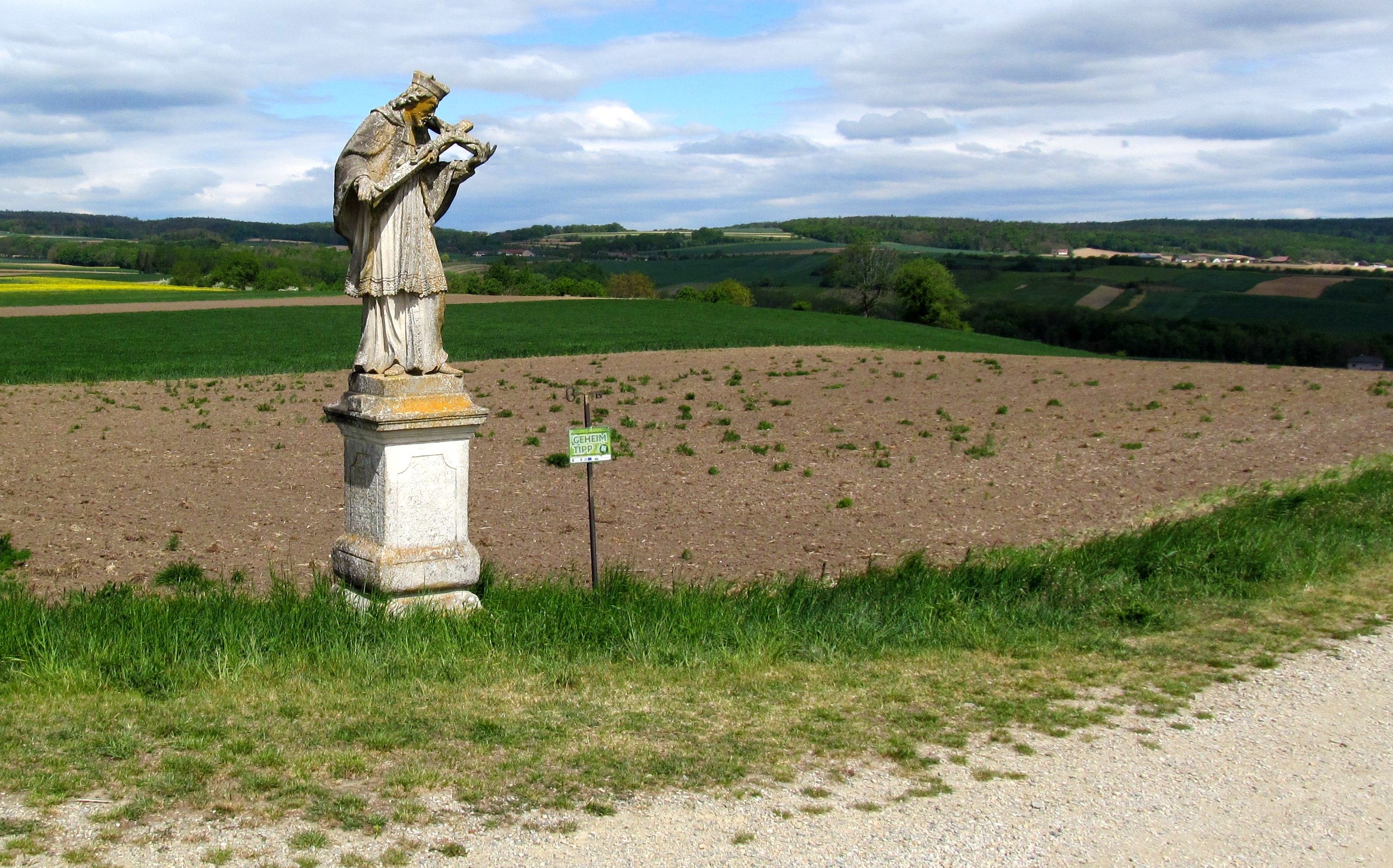 Statue of Nepomuk in a field in Baierdorf, surrounded by a green landscape and a cloudy sky.