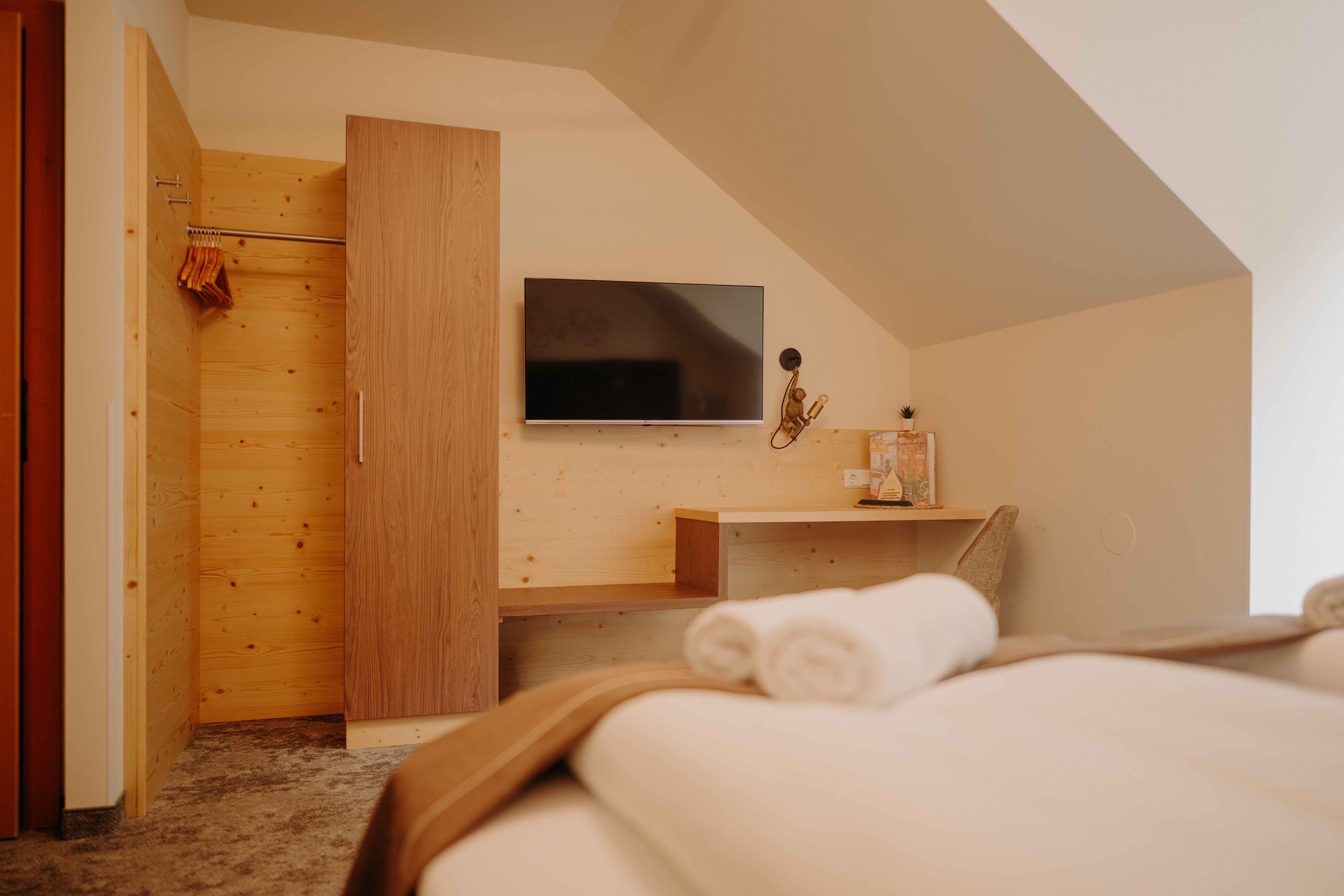 A cozy hotel room photographed from the bed. A flat-screen TV hangs on the wall across from the bed. Next to it is a light-wood wardrobe; part of the wall is also paneled with wood. There is also a shelf and a desk with a chair and a lamp. The room has a sloped ceiling.