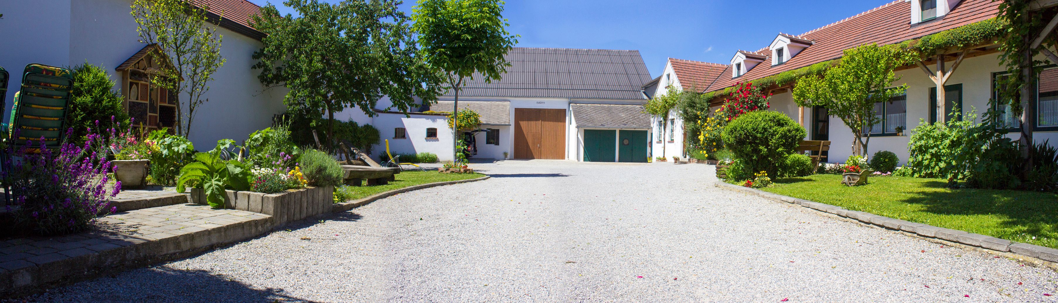 An idyllic vineyard with a gravel path, flowering gardens and traditional buildings under a blue sky.
