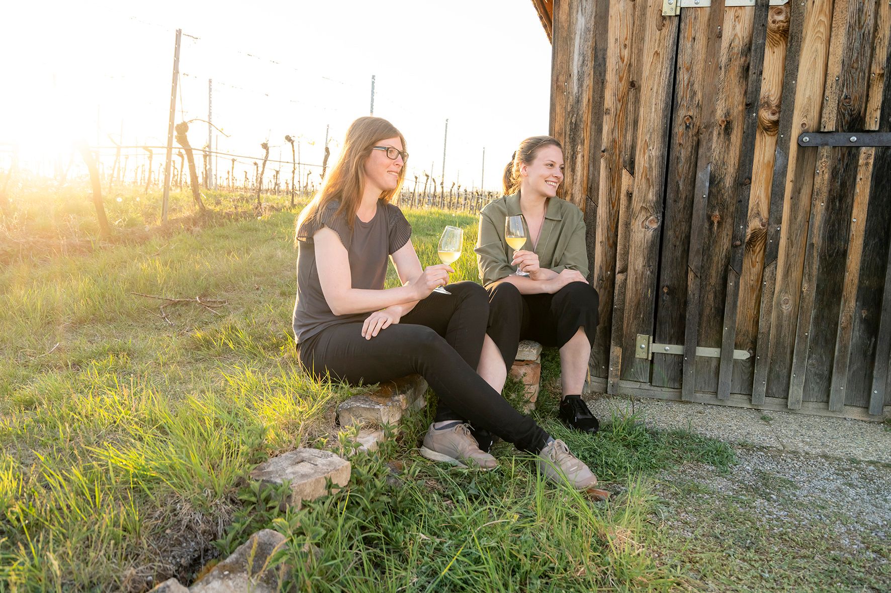 Two women sit with wine glasses in front of a wooden hut in the vineyard.