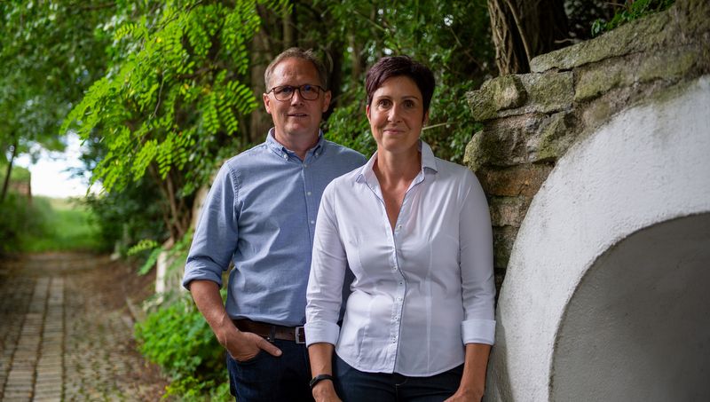 A man and a woman stand side by side on a paved path in the open air, surrounded by trees and a stone wall.