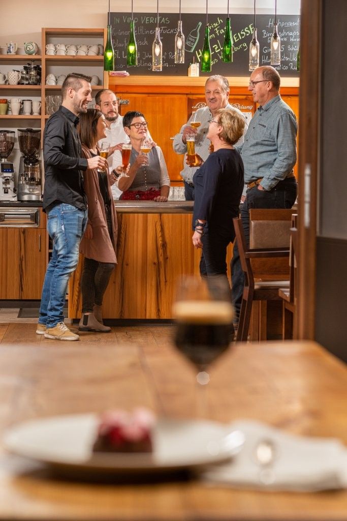 A group of people are standing in a bar holding glasses of beer. A glass and a dessert can be seen on a table in the foreground.