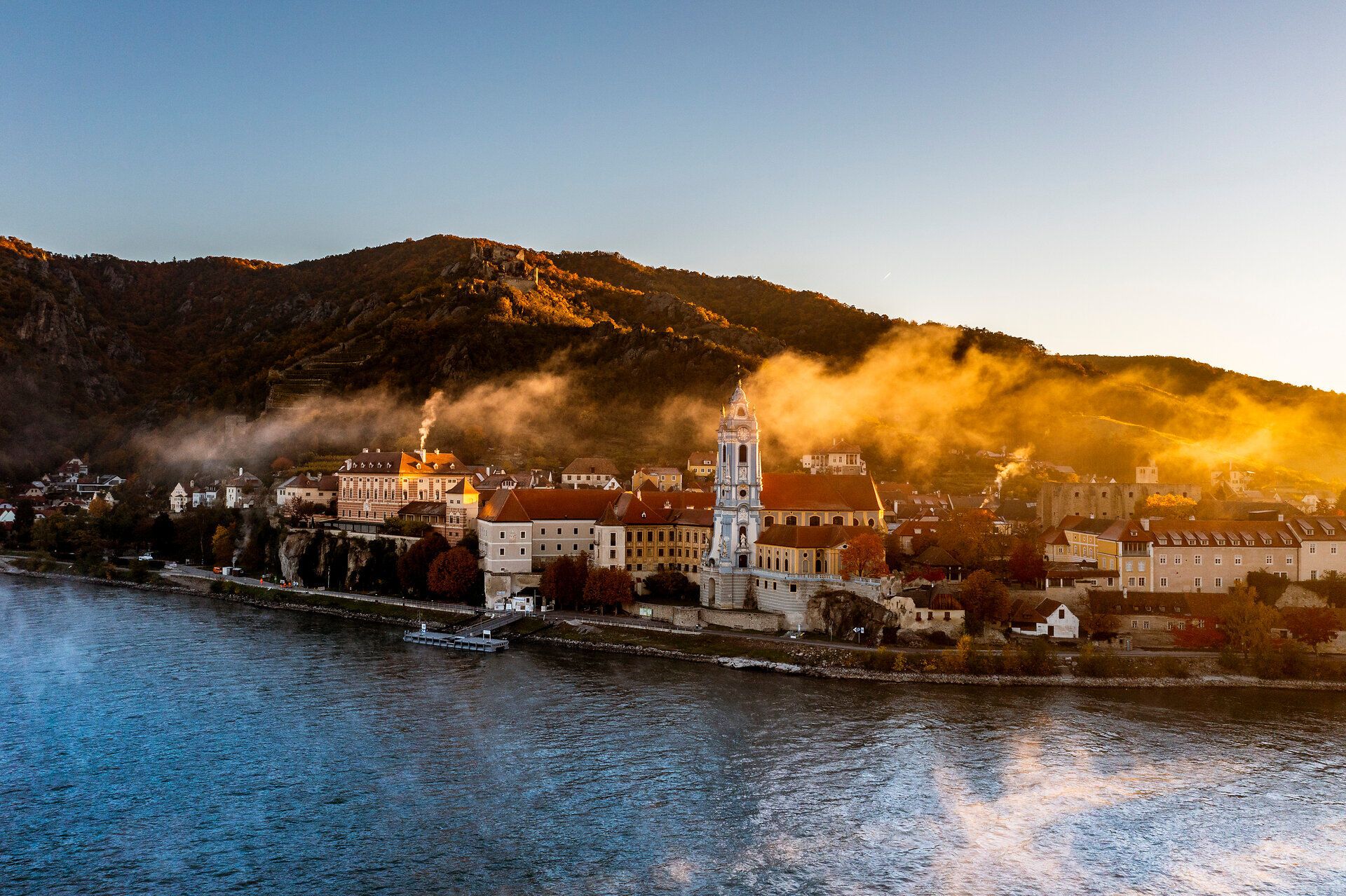 Die sanften Hügel der Wachau umarmen das malerische Stift Dürnstein, während die goldenen Strahlen der Herbstsonne die Landschaft in ein warmes Licht tauchen. Nebelschwaden ziehen über die Donau und verleihen der Szenerie eine mystische Atmosphäre, die zum Verweilen einlädt.