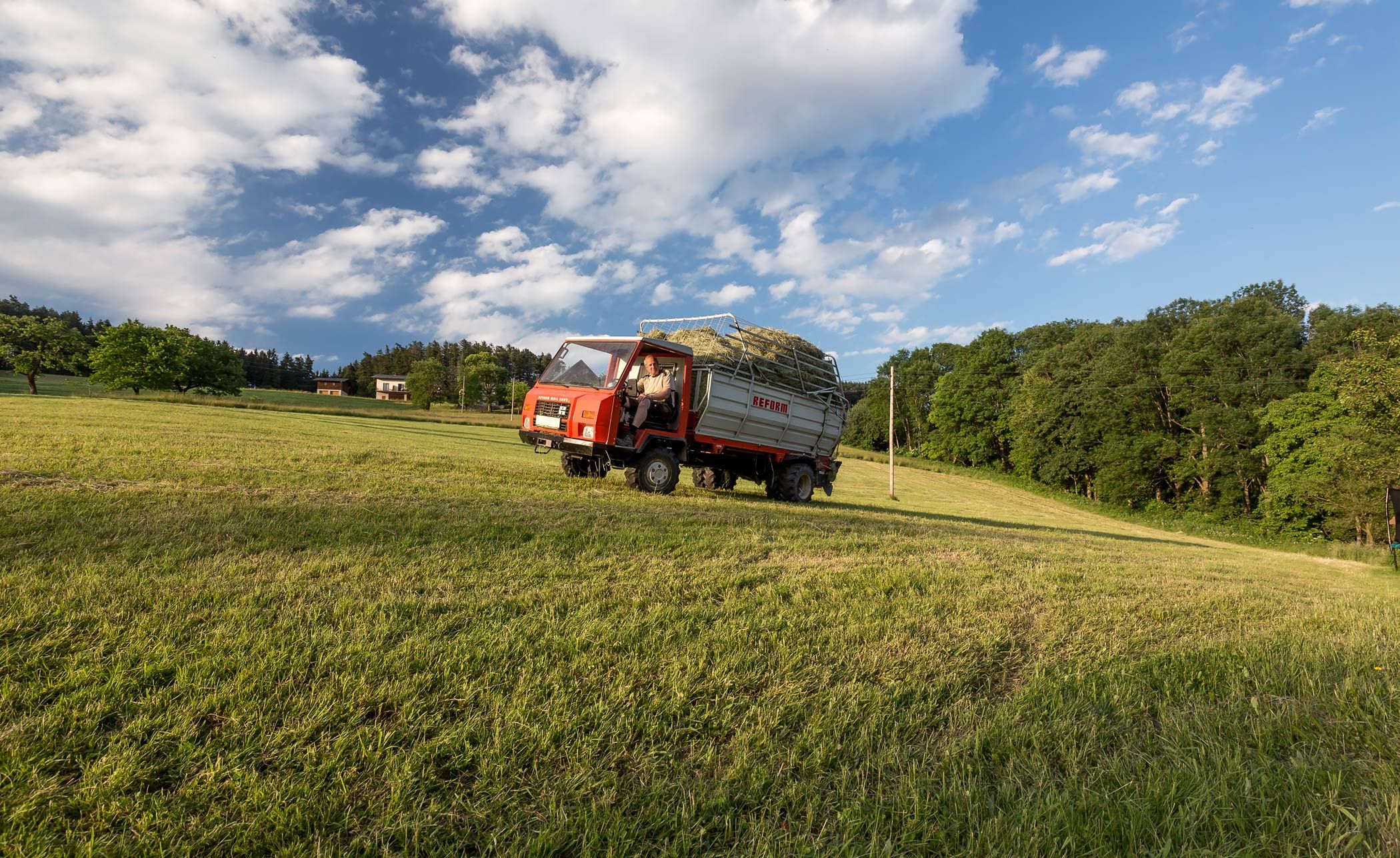 A tractor with a trailer full of hay drives across a green field under a blue sky with clouds.