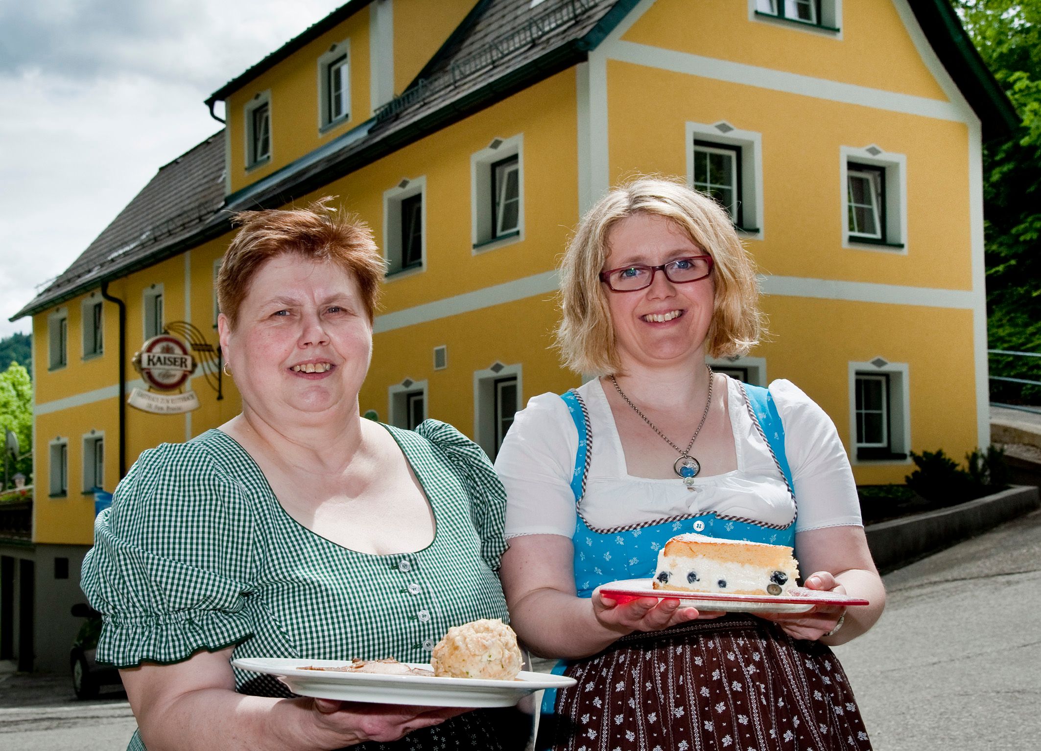 Two women in traditional dress hold plates of food in front of a yellow building.