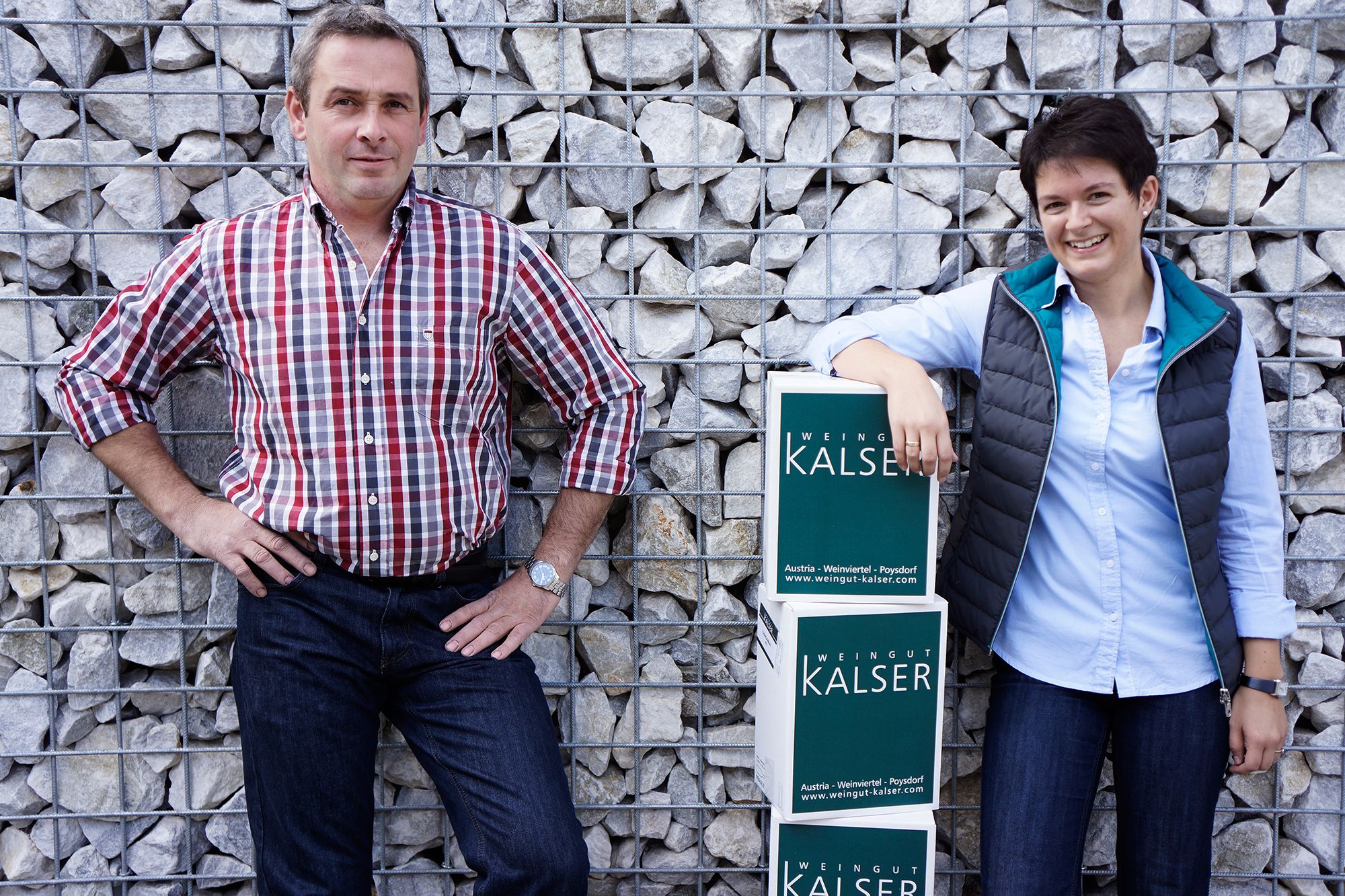 Two people are standing in front of a stone wall with crates from the Kalser winery.
