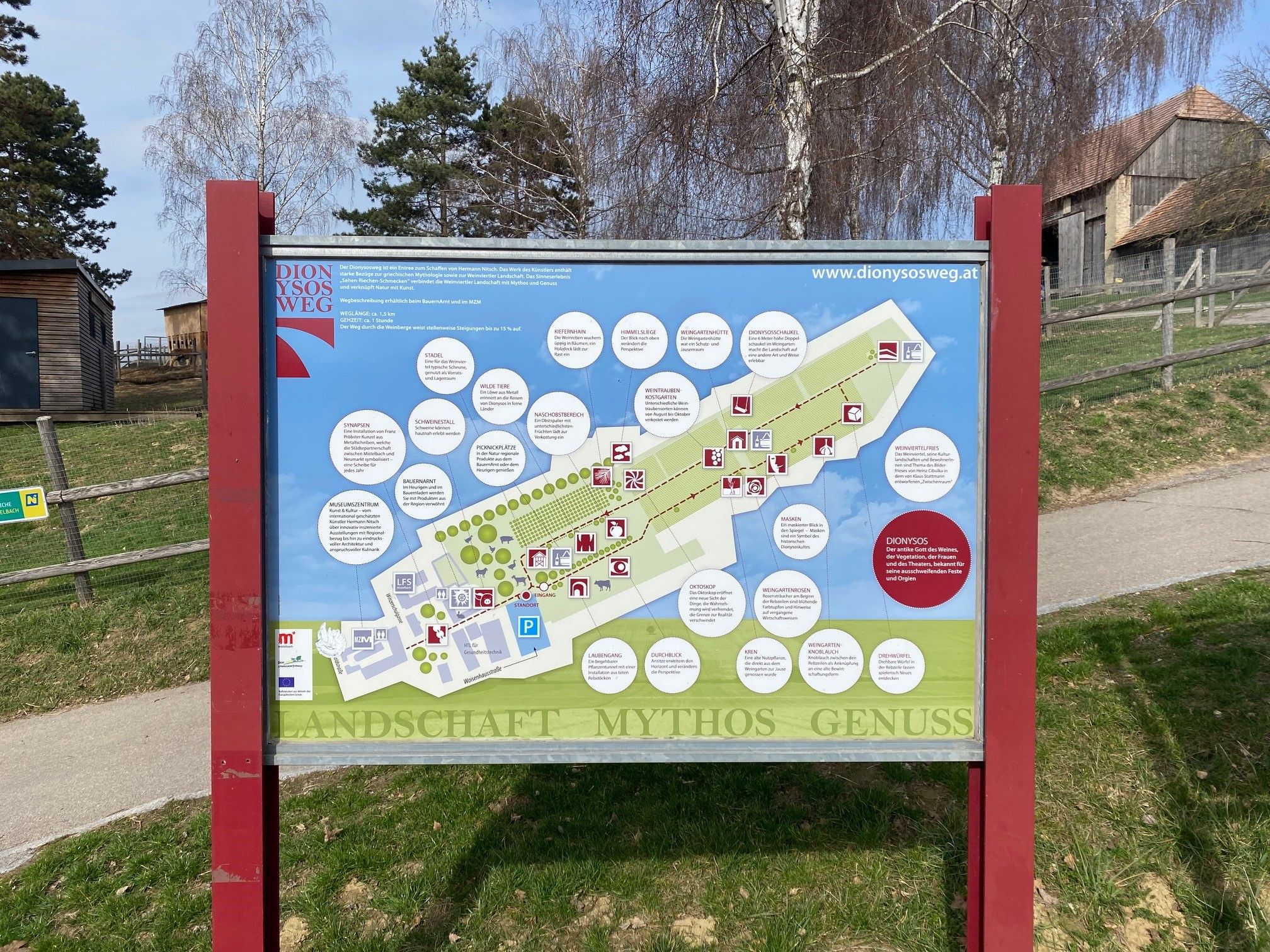 Information sign for the Dionysus Trail with map and explanations, surrounded by trees and buildings.