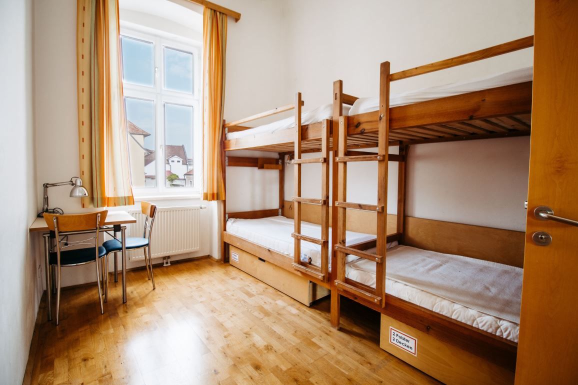 A simple room with a bunk bed, table and chair, light-colored curtains at the window.