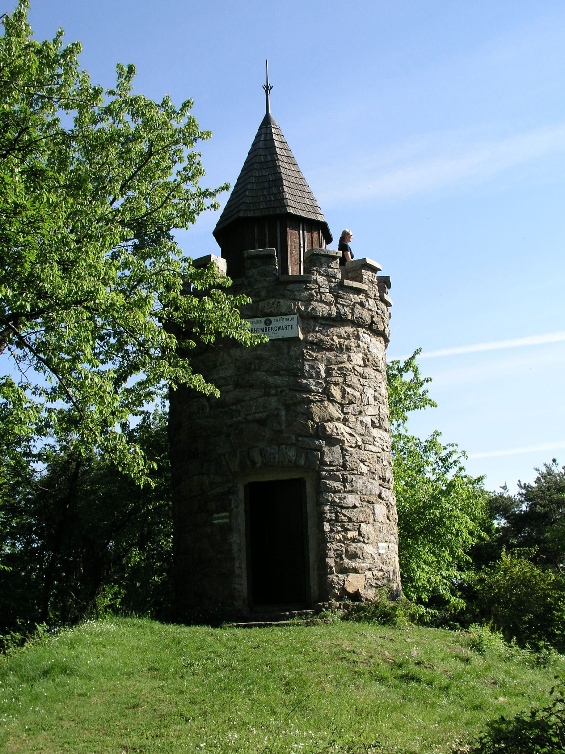 Stone observation tower with a pointed roof, surrounded by trees, under a clear sky.