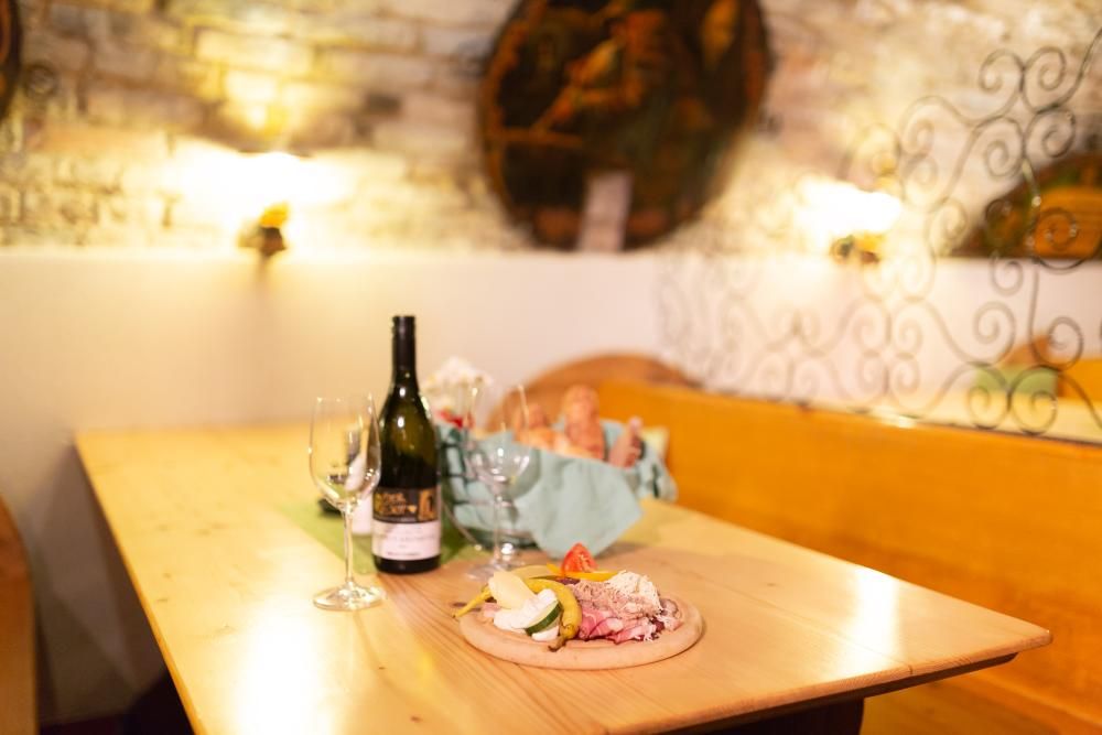 A table in a rustic room with a bottle of wine, glasses and a snack with bread and cold cuts.