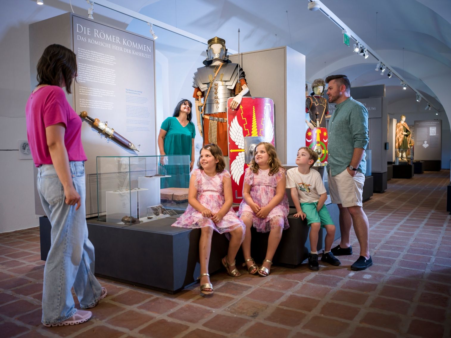 Children are sitting in front of Roman armor at the City Museum; adults are standing next to it