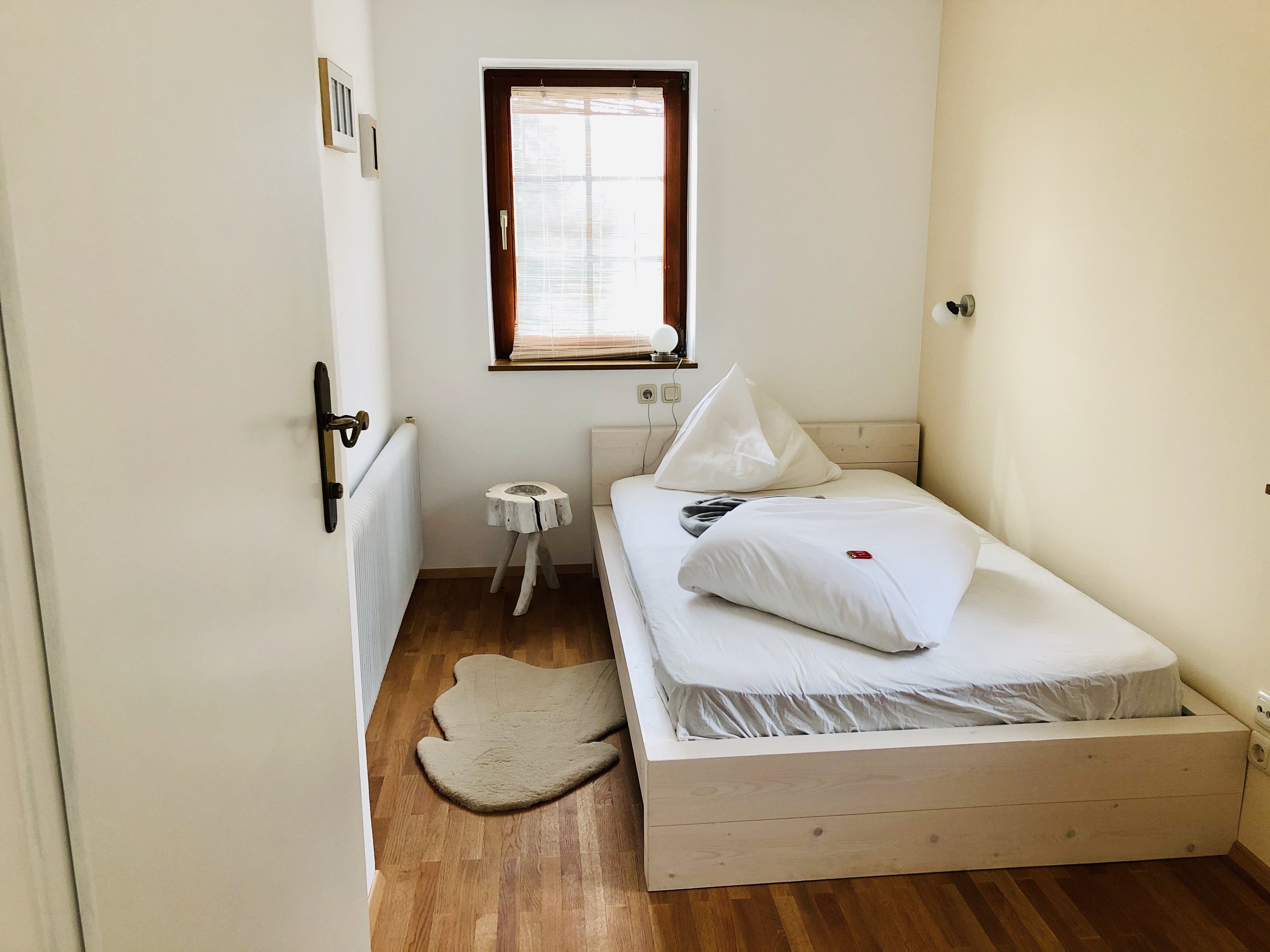 A small, cozy bedroom with a single bed, wooden floor and a small window with blinds.
