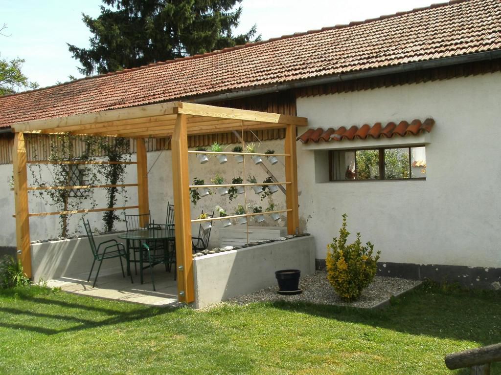 A garden with a wooden pergola, including a table and chairs. In the background a building with a tiled roof.