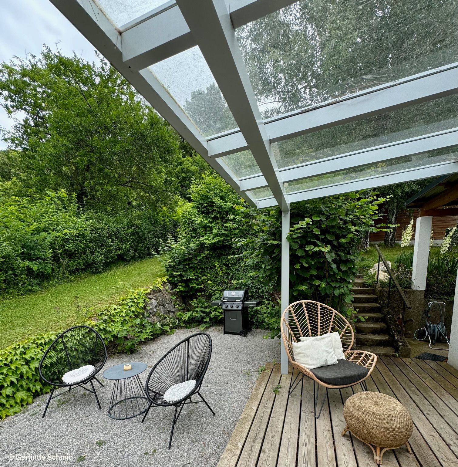 Terrace with glass roof, garden furniture and barbecue, surrounded by green vegetation.