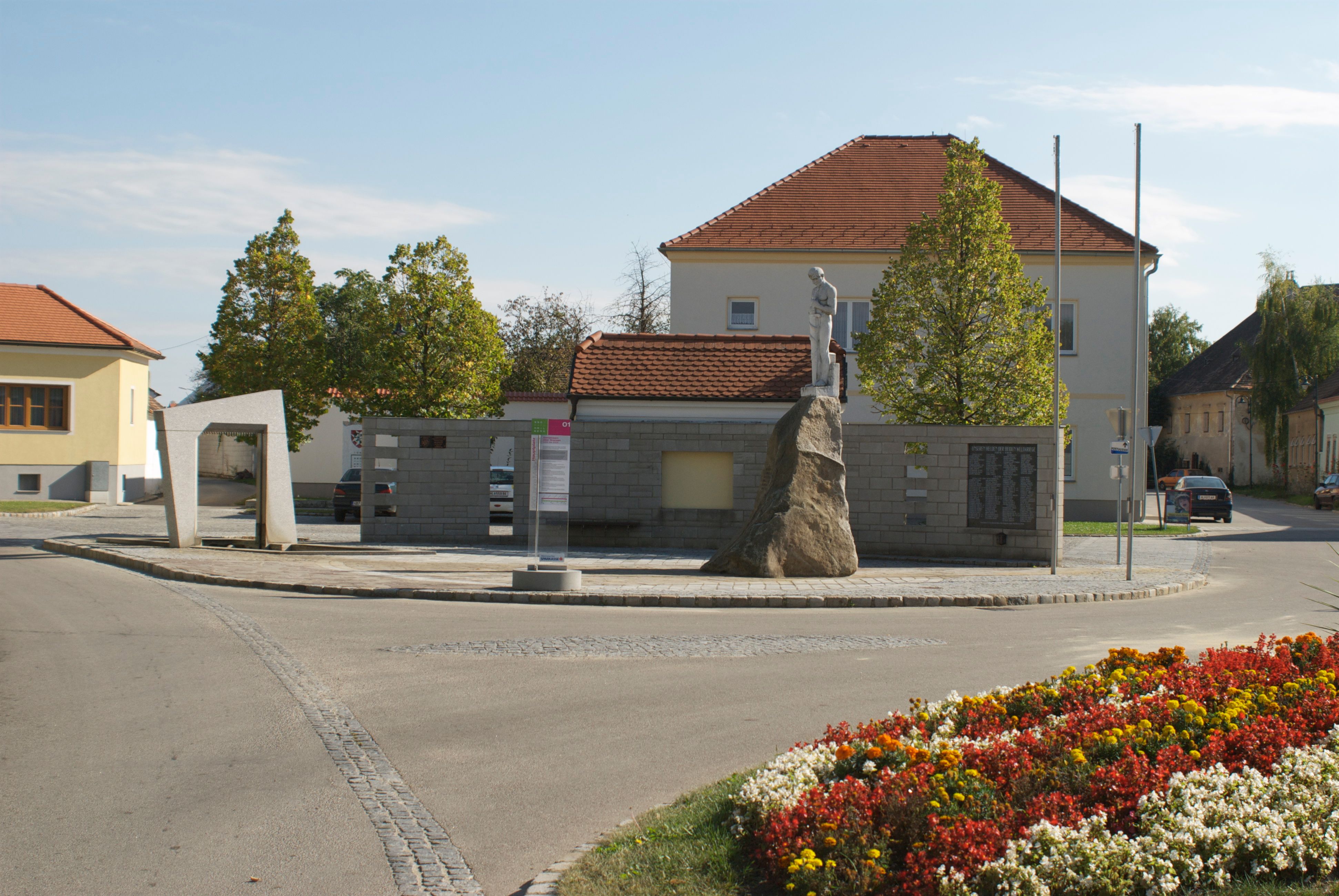 Monument in Mailberg with statue on a rock, surrounded by buildings and flowerbeds.