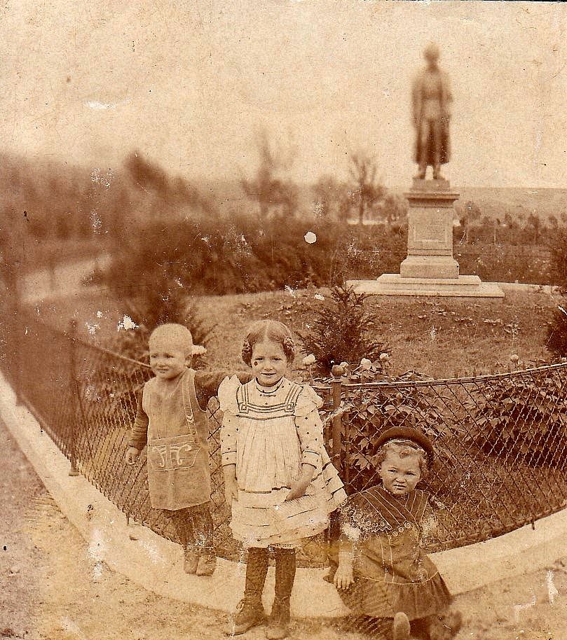 Three children are standing in front of a statue in a historic park.