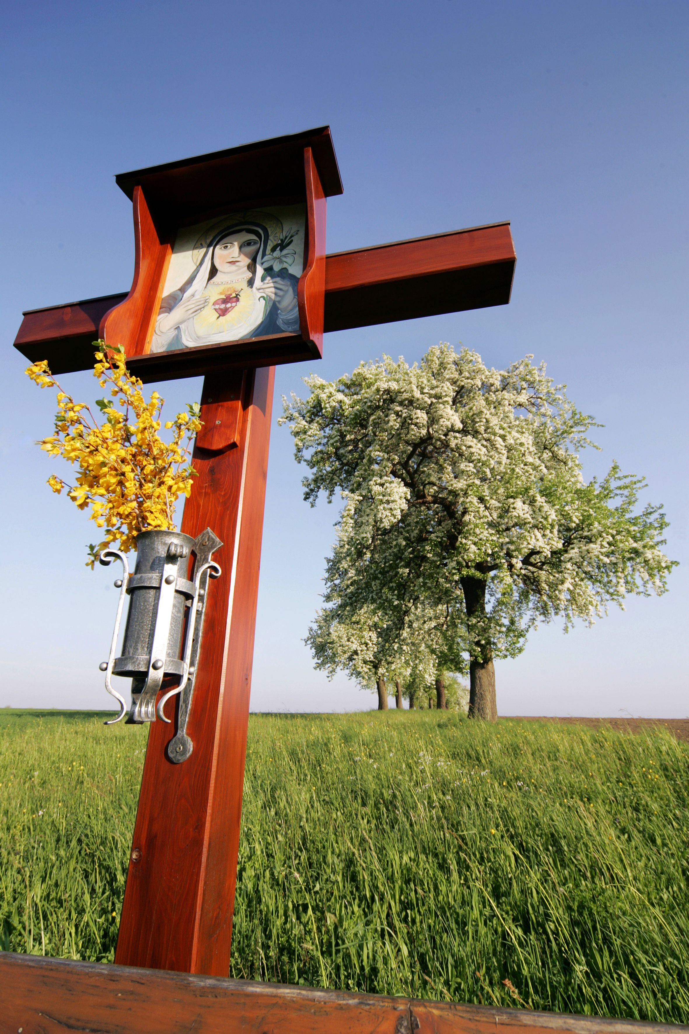 A wooden wayside cross with a picture of the Virgin Mary and yellow flowers, with blossoming trees in a meadow in the background.
