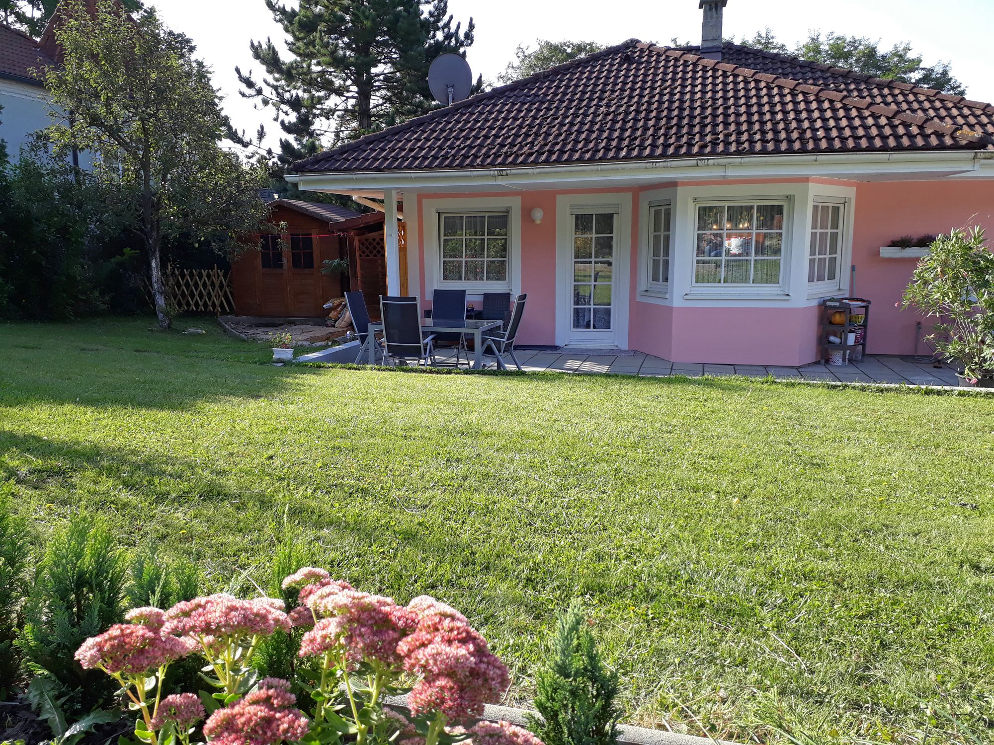 A pink house with a garden, terrace and garden furniture, surrounded by trees and flowers.