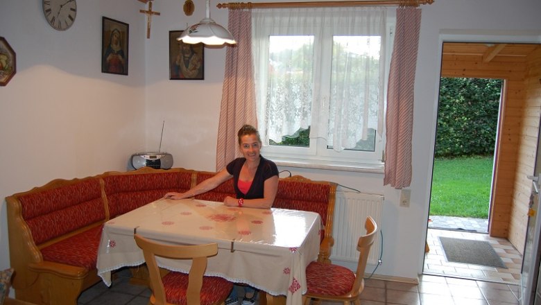 A woman sits at a dining table in a cozy kitchen with a corner bench and red cushions.