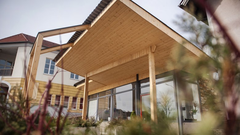 Modern building with wooden roof and glass front, surrounded by plants.