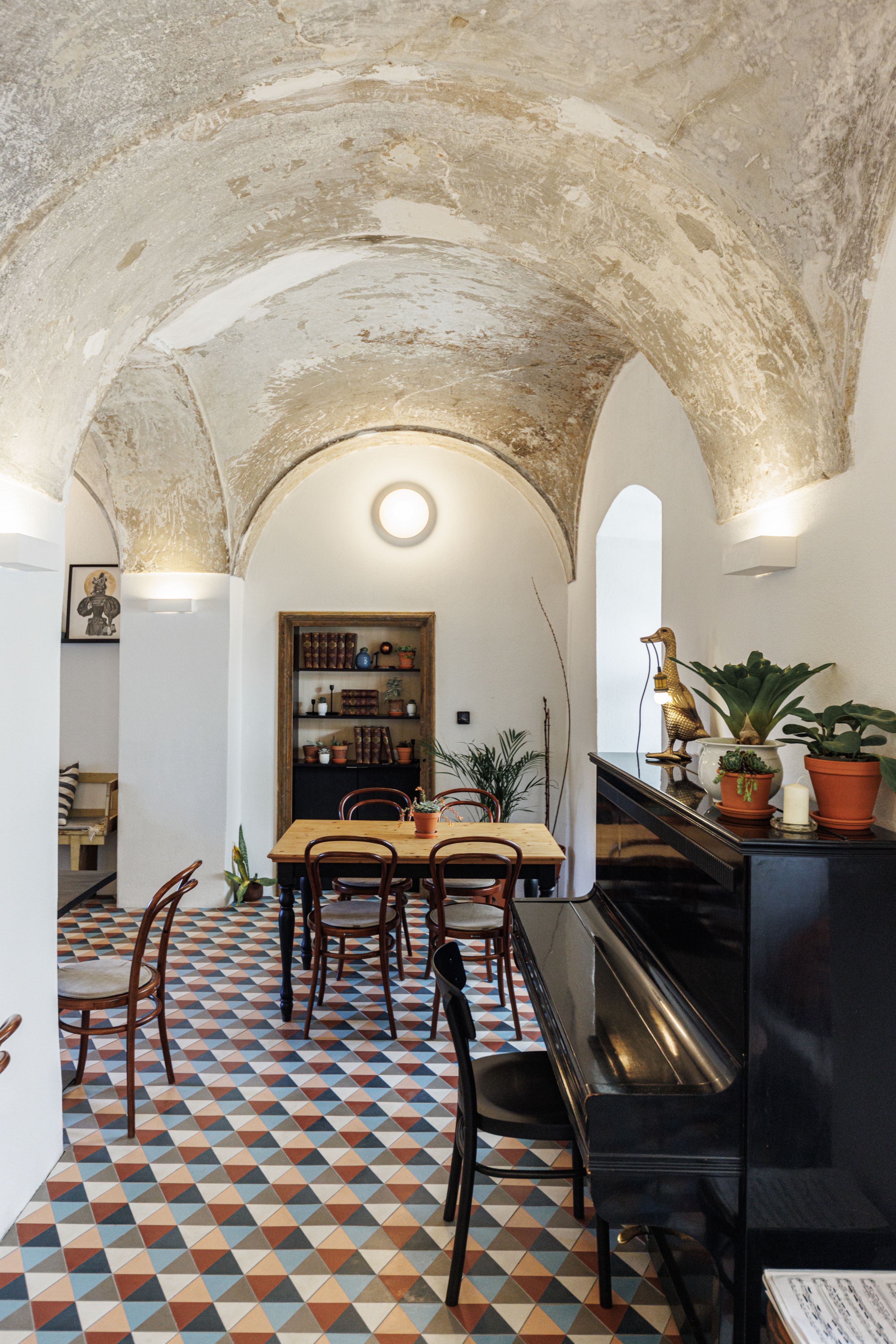 A stylish café with a vaulted ceiling, colorful tiled floor, wooden table and chairs. A piano is on the right, plants and decorations are scattered around.