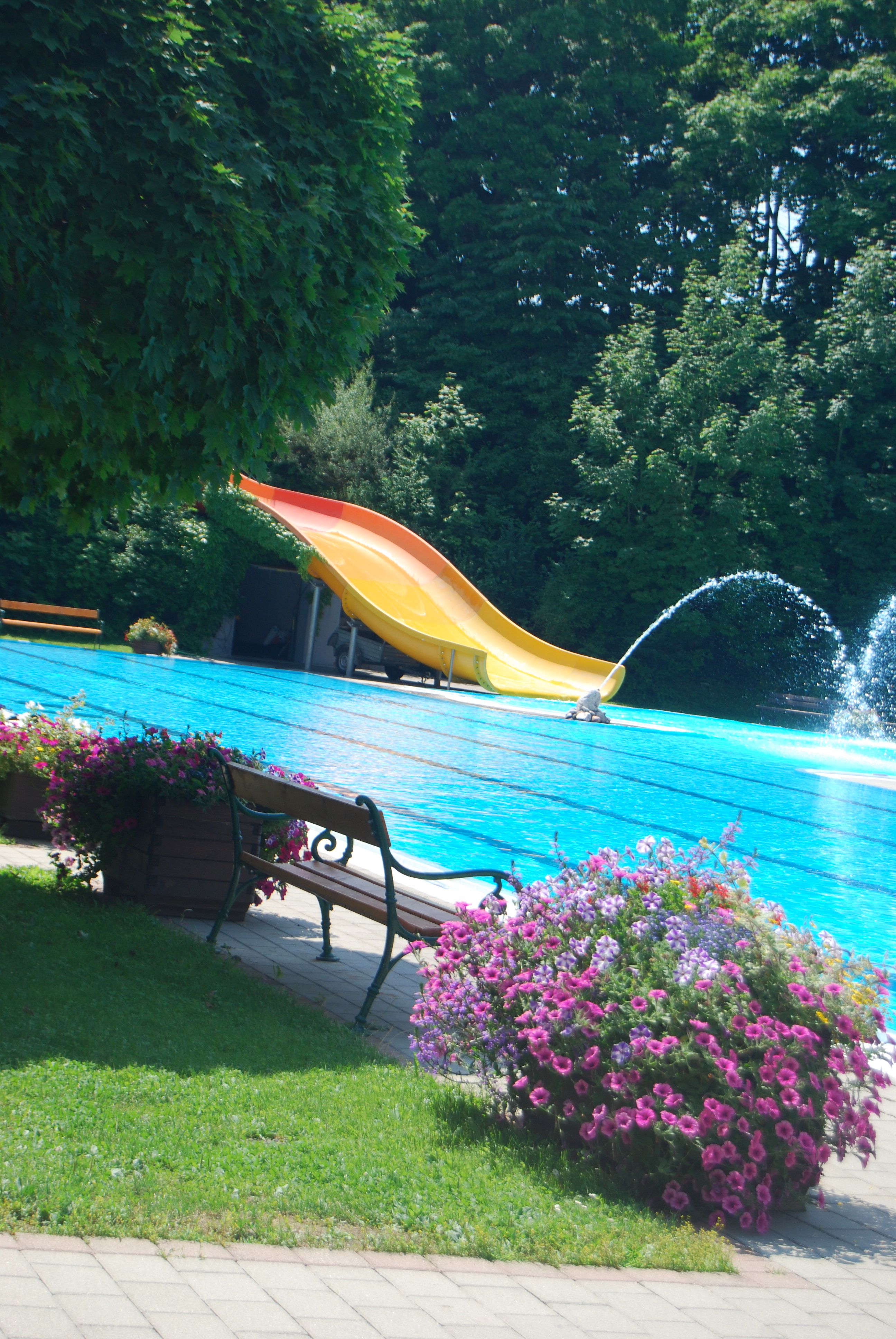 An outdoor pool with a yellow slide, water fountain and flowers in the foreground.