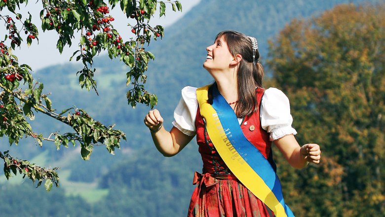 Woman in traditional costume stands under a tree with red fruits in a rural landscape.