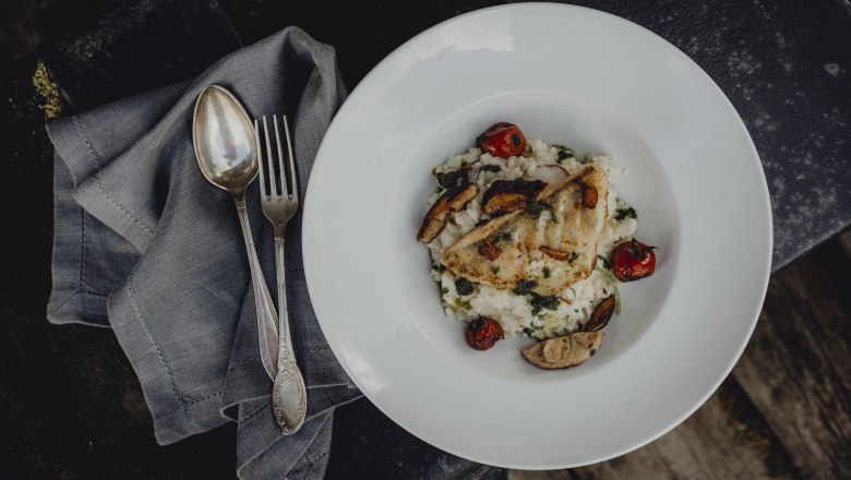 A plate with a fish dish, served on risotto with tomatoes and mushrooms, cutlery on a napkin next to it.
