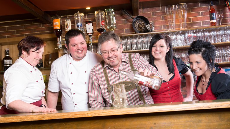 A group of people behind a bar, laughing together and pouring a bottle into a jug.