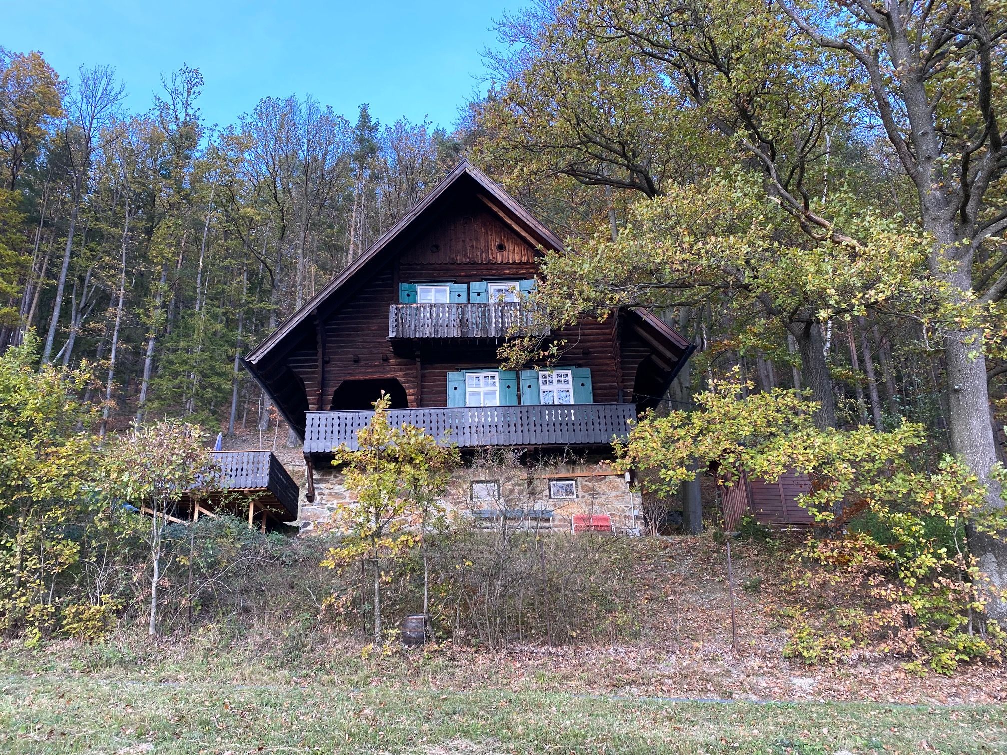 A rustic wooden house with a balcony stands on the edge of the forest, surrounded by trees and autumn leaves.