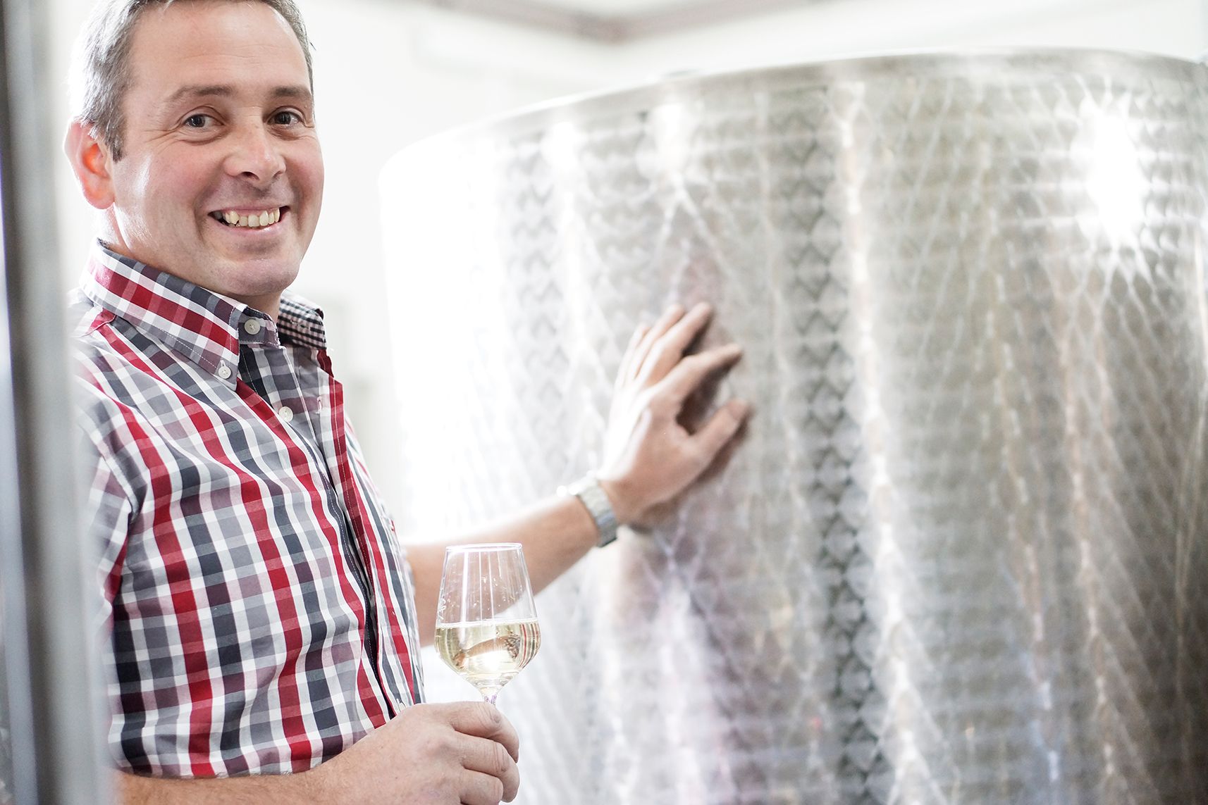Man in plaid shirt holding a wine glass next to a large metal tank.