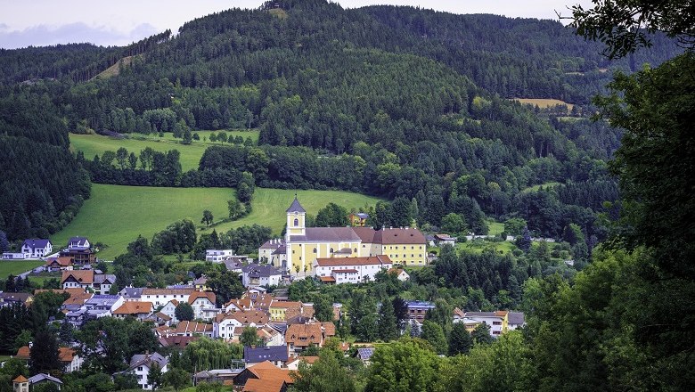 A church towers over a picturesque village, surrounded by green hills and forests.