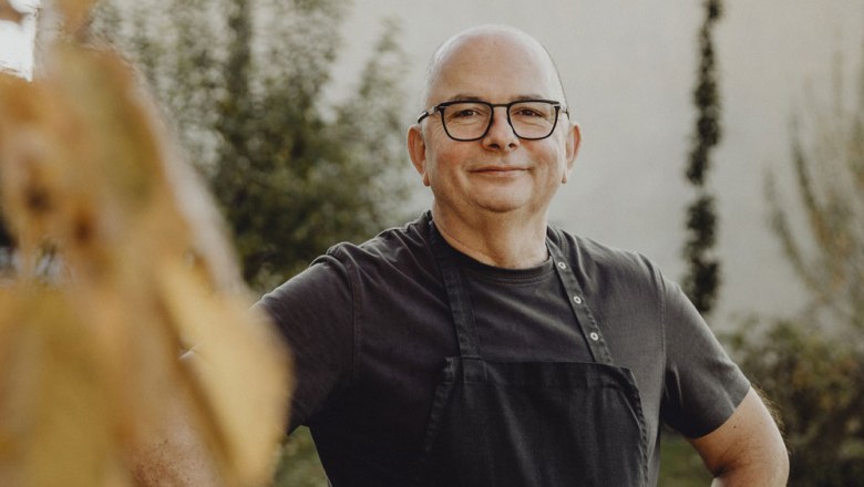 Man with glasses and apron smiling outdoors, blurred background.
