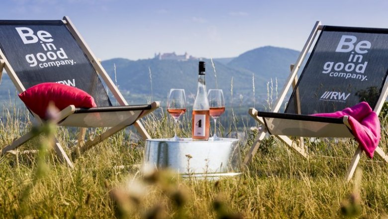 Two deckchairs with the inscription 'Be in good company' stand on a lawn. Between them is a table with a bottle of wine and two glasses. Mountains in the background.