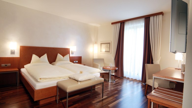 A modern hotel room with double bed, wooden floor, desk and large window with curtains.