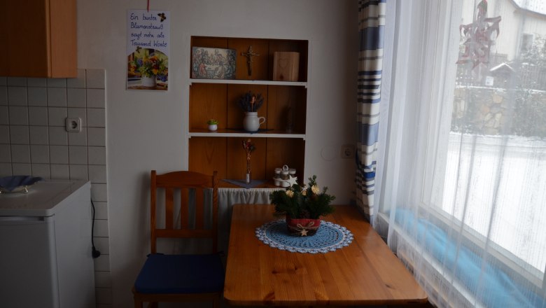 Small kitchen with wooden table, chair and shelf. Window with curtain, snow outside. Decorations and plants on table and shelf.