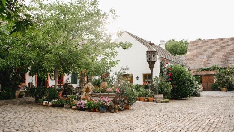 An idyllic courtyard with cobblestones, surrounded by green trees and colorful flowers, in front of a white building with a tiled roof.