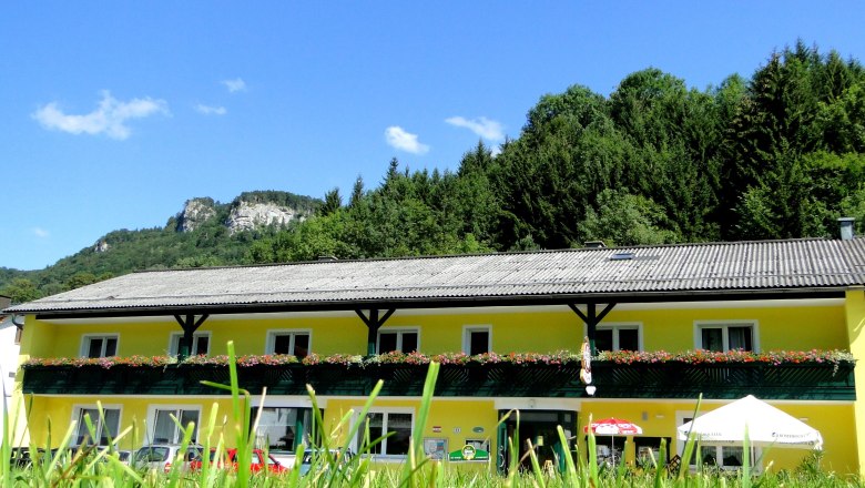 Yellow building with flower balcony in front of a wooded hill and blue sky.