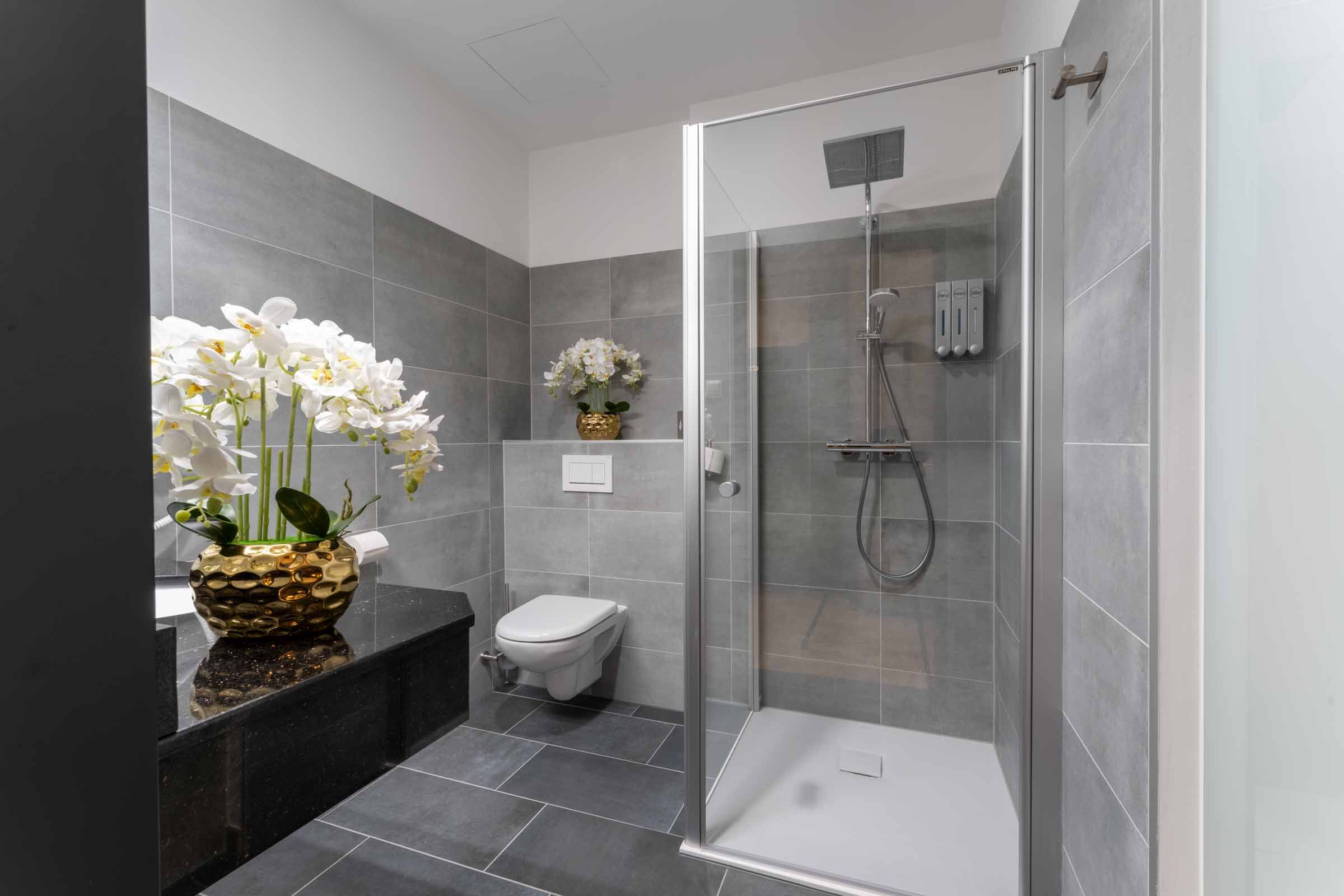 Modern bathroom with gray tiles, shower, toilet and orchids in golden vases.