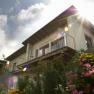 Exterior view of a house with balcony and flowering garden in the foreground, rays of sunshine in the sky.