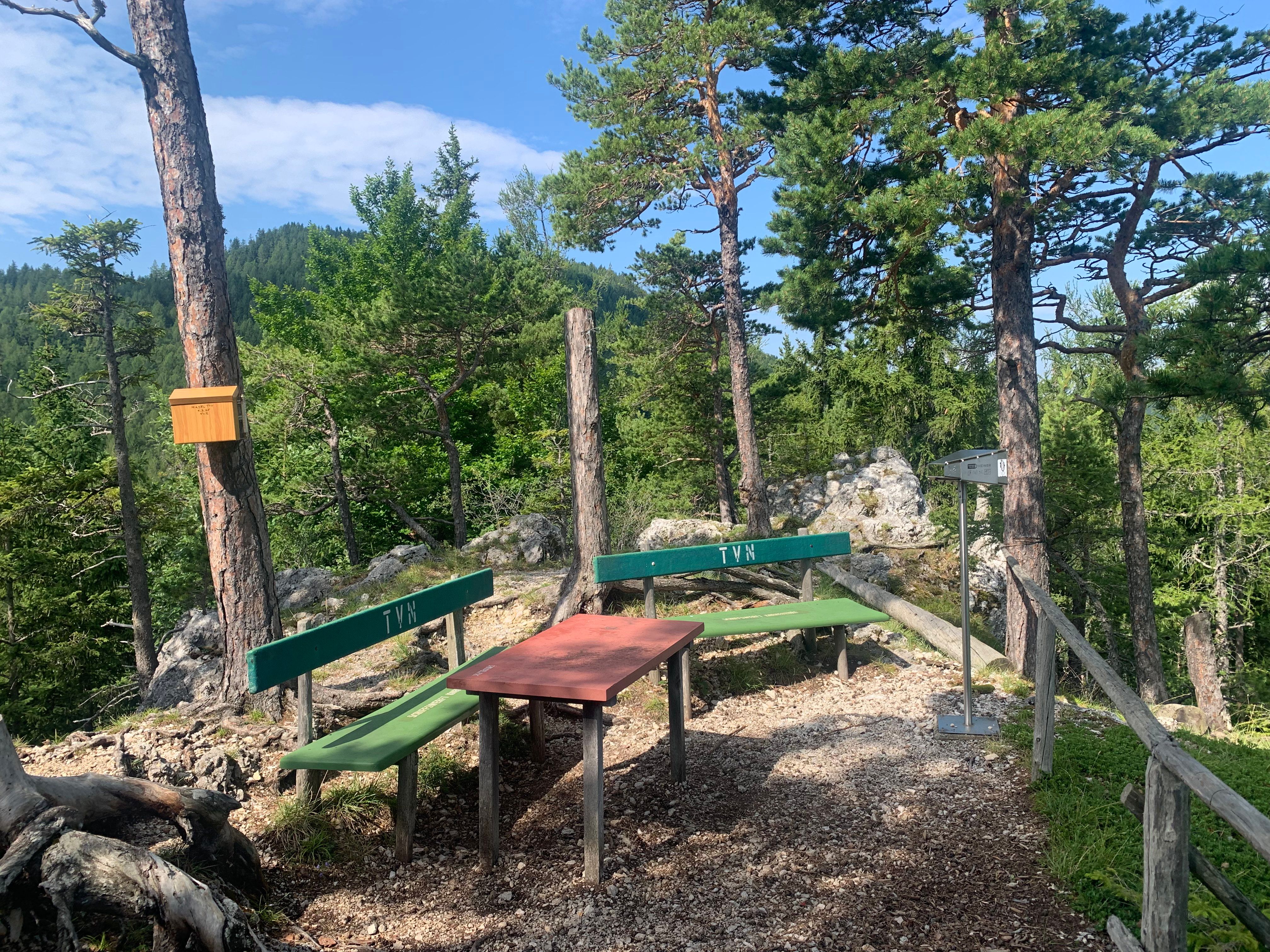 A rest area in the forest with two benches and a table, surrounded by trees and rocks.
