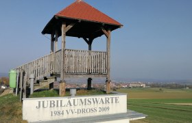 Wooden lookout with a red roof on a hill, surrounded by fields and a village in the background.