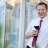 Man in white shirt and red tie holding a glass of red wine in front of a building.