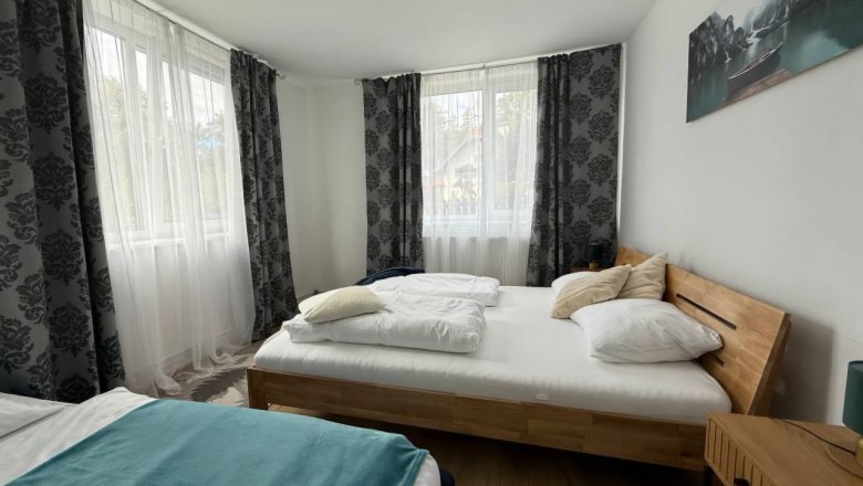 Bright bedroom with two beds, wooden floor and large windows with curtains.